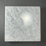 Square translucent slice of selenite crystal with a textured, faceted pattern and white streaks on a dark surface.
