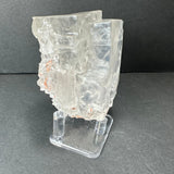 Translucent cubic fluorite crystal with irregular edges and subtle internal fractures, mounted on a clear acrylic stand against a dark gray background.