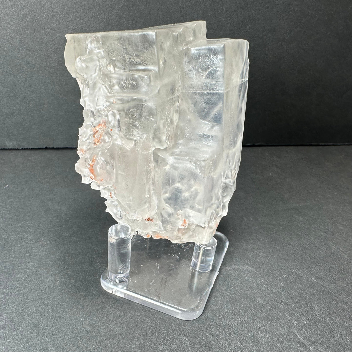 Translucent cubic fluorite crystal with irregular edges and subtle internal fractures, mounted on a clear acrylic stand against a dark gray background.