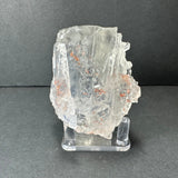 Colorless cubic fluorite crystal from Hardin County, Illinois, with transparent facets and small reddish inclusions, displayed on a clear stand against a gray background.