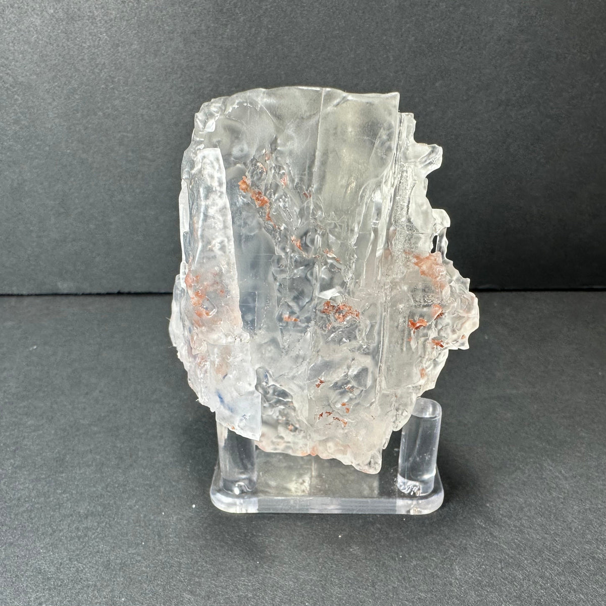 Colorless cubic fluorite crystal from Hardin County, Illinois, with transparent facets and small reddish inclusions, displayed on a clear stand against a gray background.