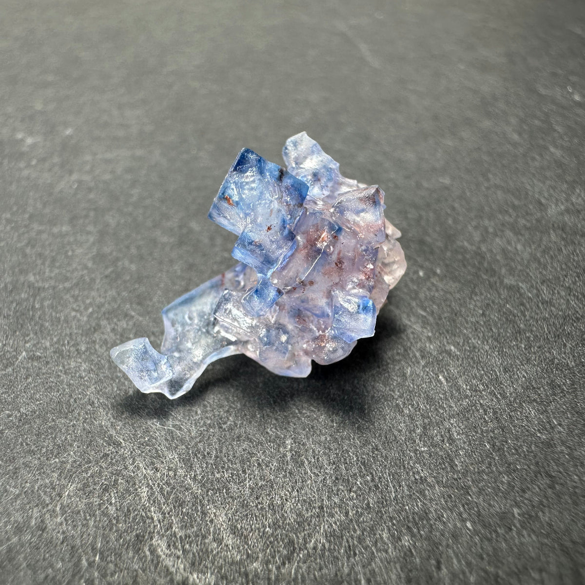 Blue fluorite crystal cluster with cubic shapes and translucent light purple and blue tones on a gray surface.