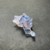 Cluster of translucent blue fluorite cubes with subtle purple and white hues, resting on a textured dark gray surface.