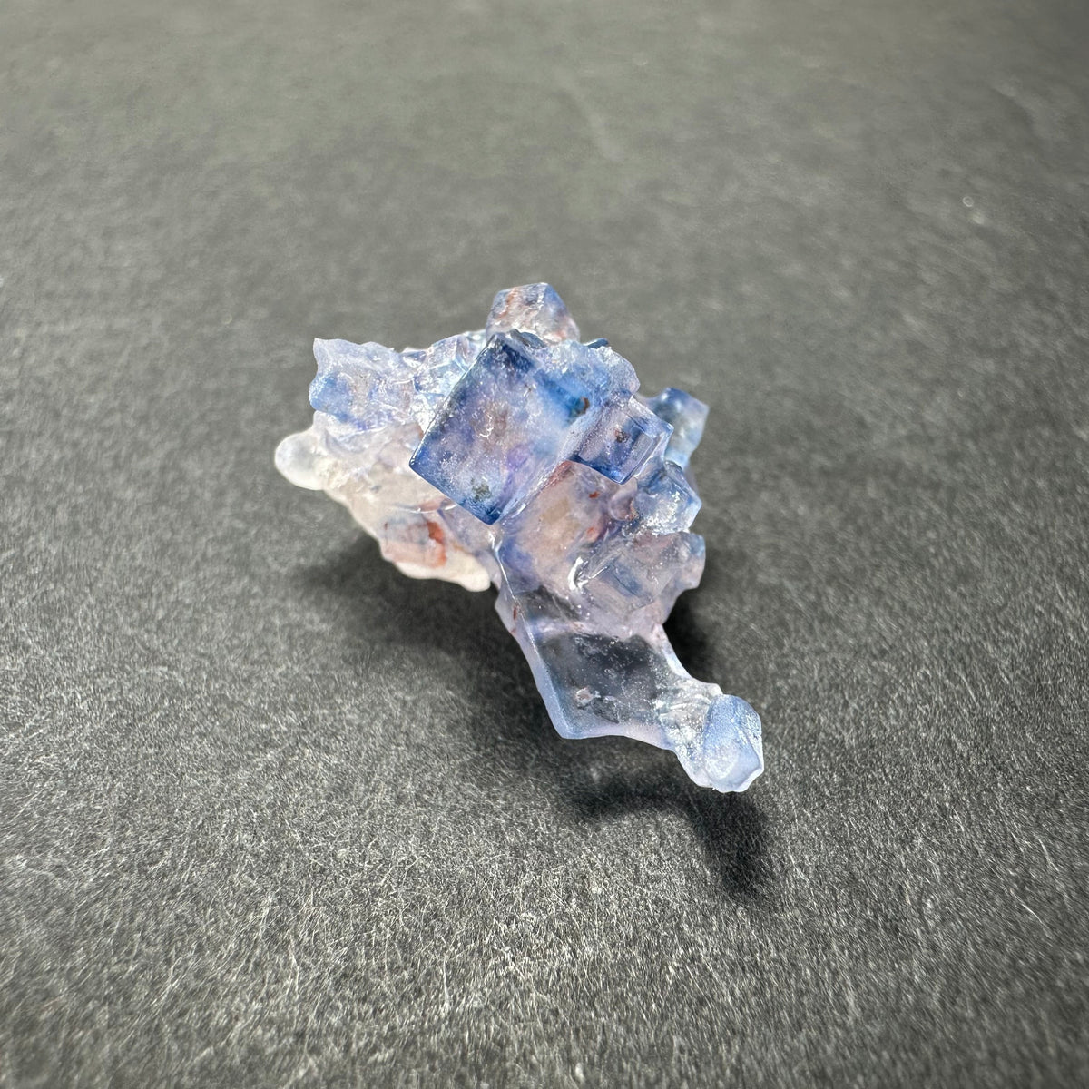 Cluster of translucent blue fluorite cubes with subtle purple and white hues, resting on a textured dark gray surface.