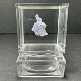 Small blue fluorite cube with purple hues displayed suspended inside a clear acrylic cube case with a matching base, against a dark gray background.