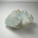 Translucent pale blue aquamarine crystal cluster with angular, well-defined shapes and smaller white crystalline formations attached, shown on a plain white surface.