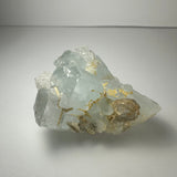 Translucent aquamarine crystal cluster with pale blue hues and golden streaks, showing angular facets and an irregular natural form, set against a neutral background.