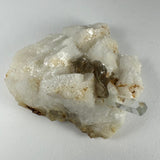 White, blocky Albite mineral with a transparent prism-shaped Aquamarine crystal and dark brown layered Muscovite on top, displayed on a gray background.