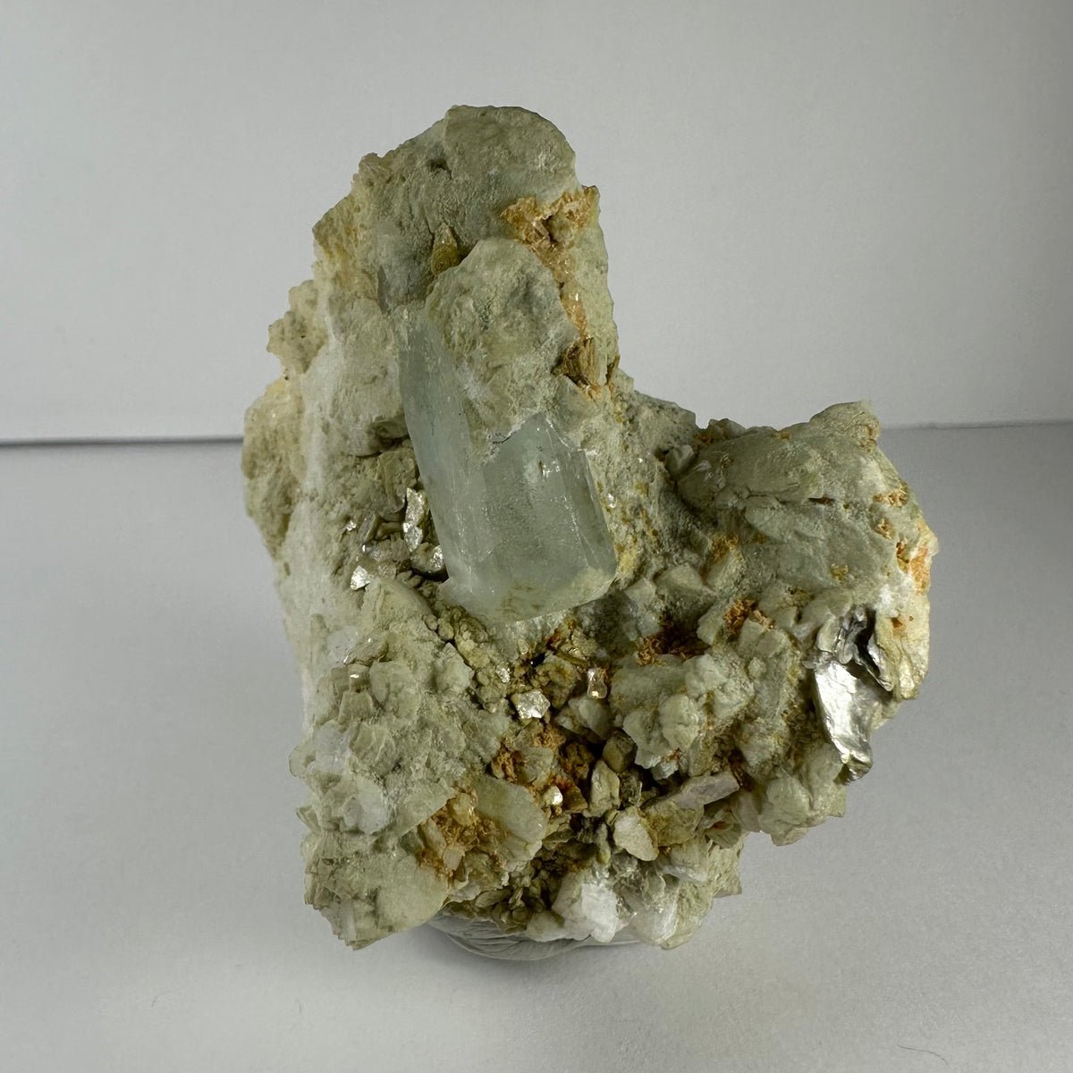 Pale aquamarine crystal embedded in a rough beige feldspar, albite, and mica matrix with reflective metallic mica flakes, displayed on a clear stand.