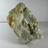Rough pale green aquamarine crystal embedded in a white and beige feldspar, albite, and mica matrix with crystalline formations, set against a plain background.