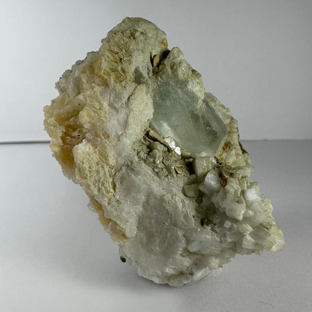 Rough pale green aquamarine crystal embedded in a white and beige feldspar, albite, and mica matrix with crystalline formations, set against a plain background.