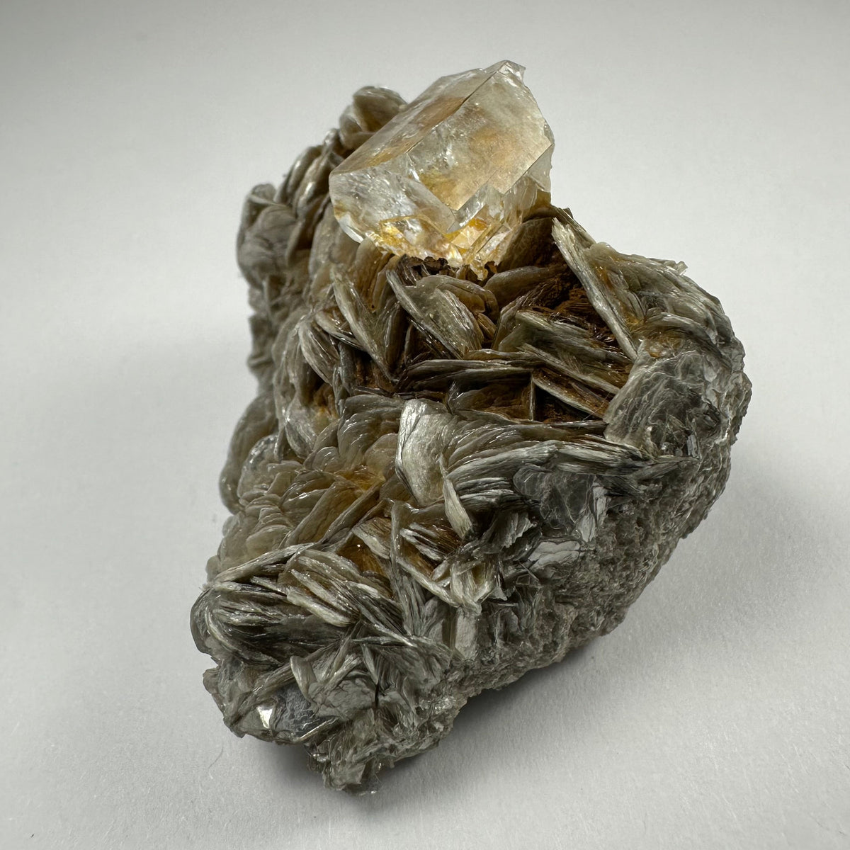 Transparent aquamarine crystal atop layered, silvery muscovite mineral matrix with intricate, curved, flaky formations, displayed on a plain light gray background.