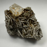 Transparent aquamarine crystal resting atop layered silvery-gray muscovite flakes with a textured, irregular shape against a neutral background.