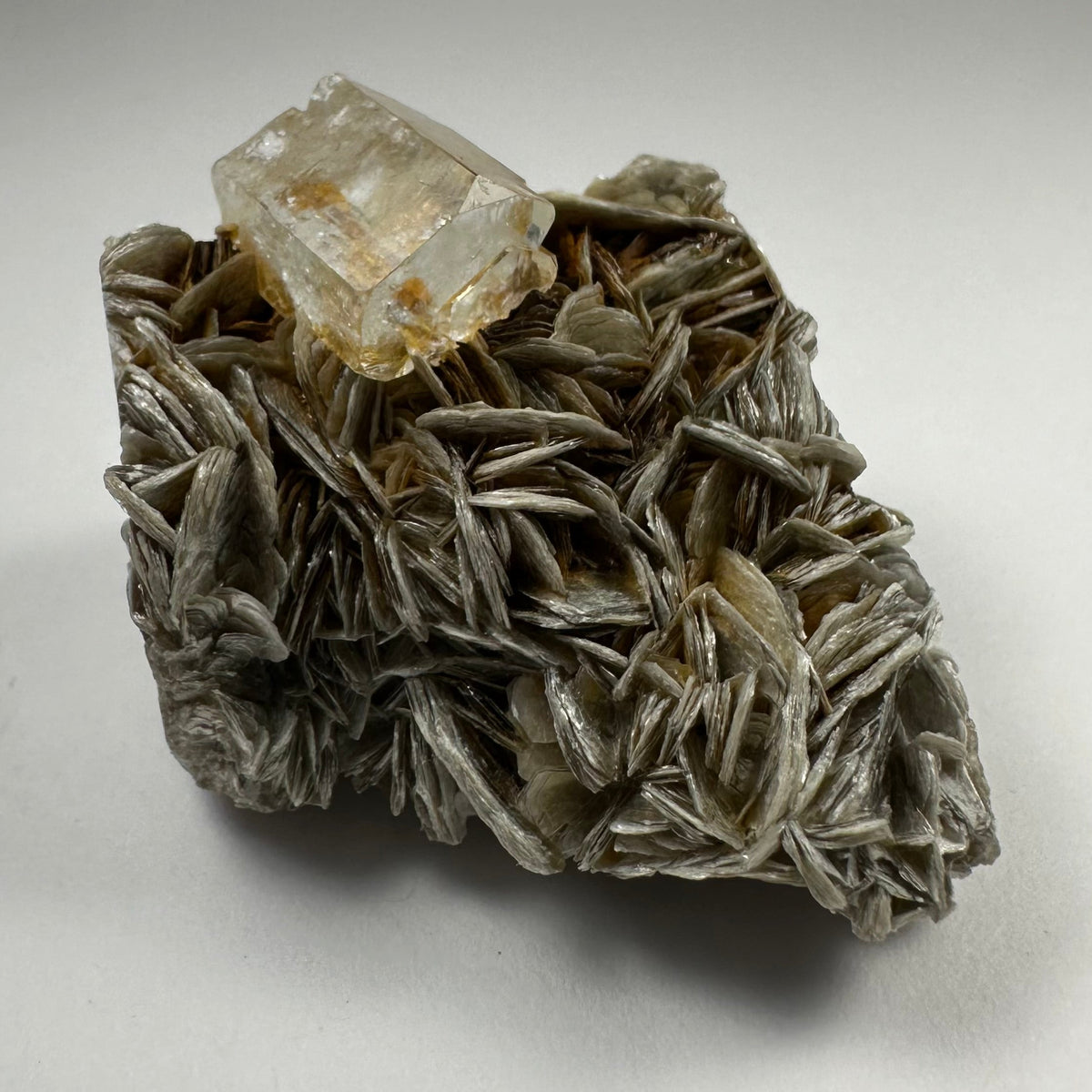 Transparent aquamarine crystal resting atop layered silvery-gray muscovite flakes with a textured, irregular shape against a neutral background.