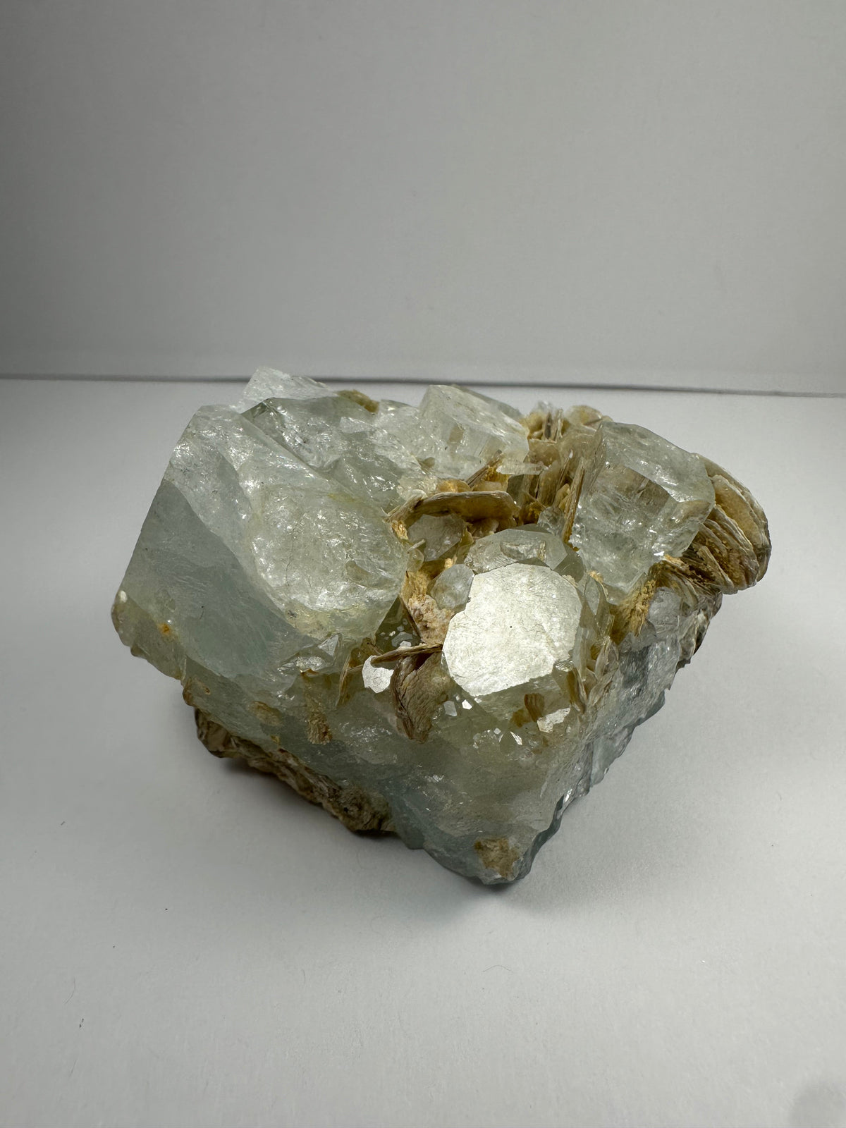 Chunk of aquamarine with translucent pale blue crystals and tan muscovite flakes clustered on top, displayed against a plain white background.