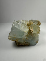 Chunky aquamarine crystal cluster with translucent pale blue color and attached beige muscovite flakes, displayed against a plain gray background.