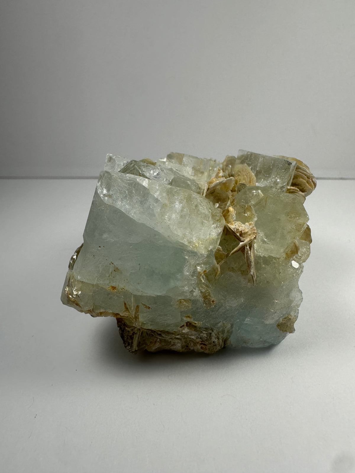 Chunky aquamarine crystal cluster with translucent pale blue color and attached beige muscovite flakes, displayed against a plain gray background.