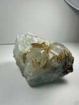 Clear aquamarine crystal with beige muscovite inclusions, showing rough, irregular surfaces and a translucent bluish tint, placed on a plain light background.
