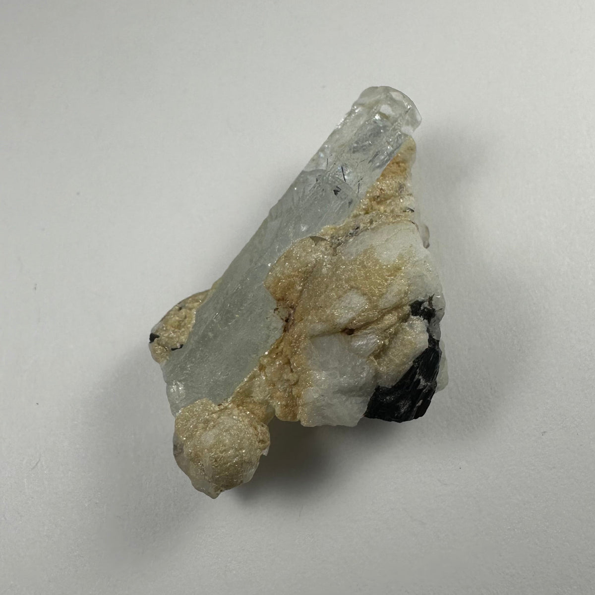 Clear aquamarine crystal embedded in white albite with tan mineral deposits and a small black tourmaline section, shown against a plain white background.