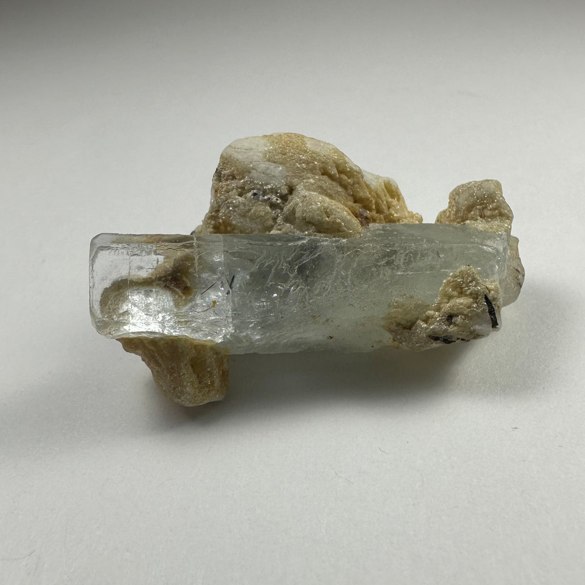 Translucent pale blue aquamarine crystal partially embedded in beige albite matrix with a small black tourmaline inclusion, shown on a plain white surface.