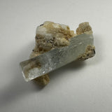 Translucent aquamarine crystal extending from beige albite matrix with small black tourmaline inclusions, displayed on plain light surface