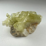 Cluster of translucent yellowish-green Brazilianite crystals with brownish and white base, displayed on a light gray surface.