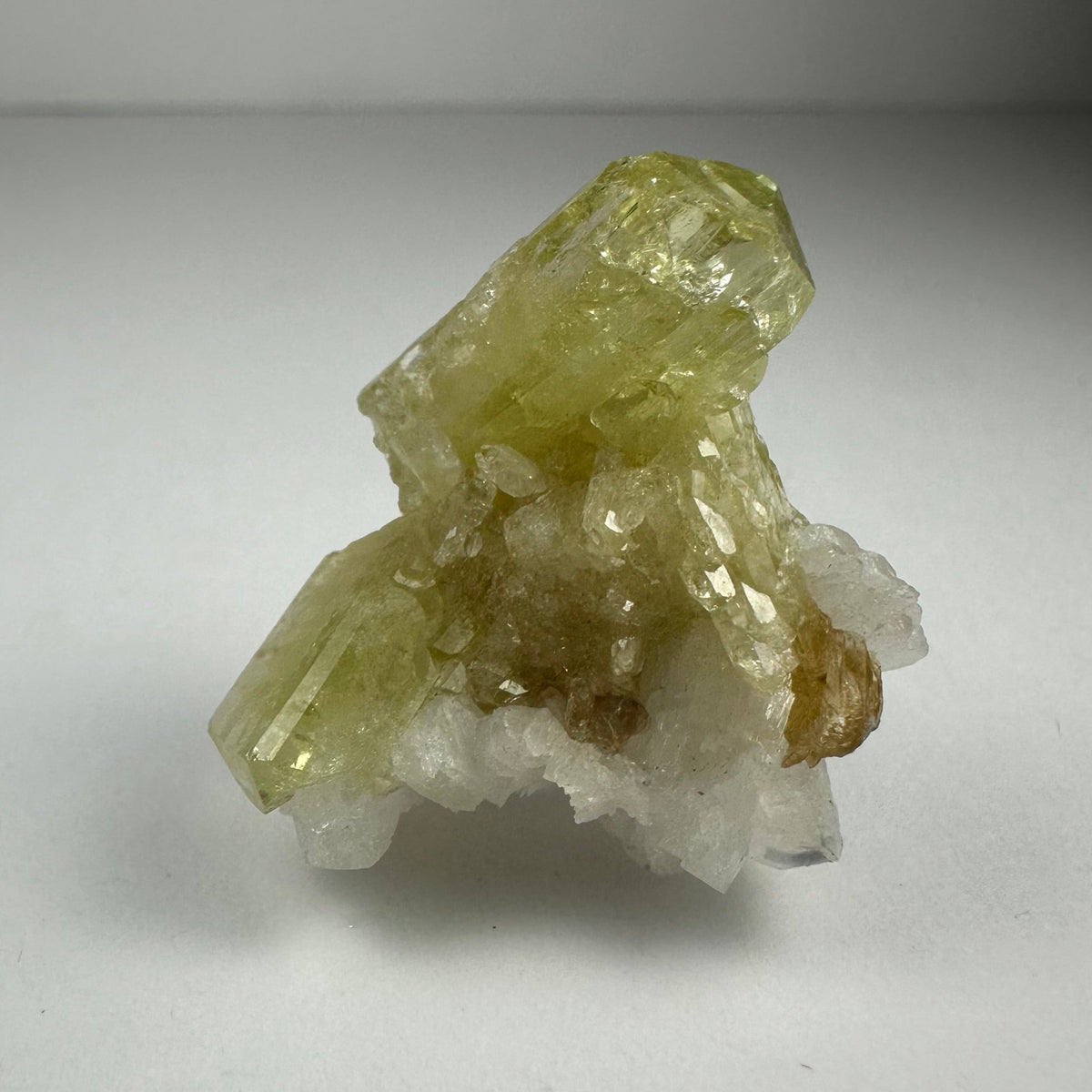 Pale yellow Brazilianite crystals with hexagonal shapes resting on white Albite matrix with small brown mineral inclusions, set against a neutral gray background.