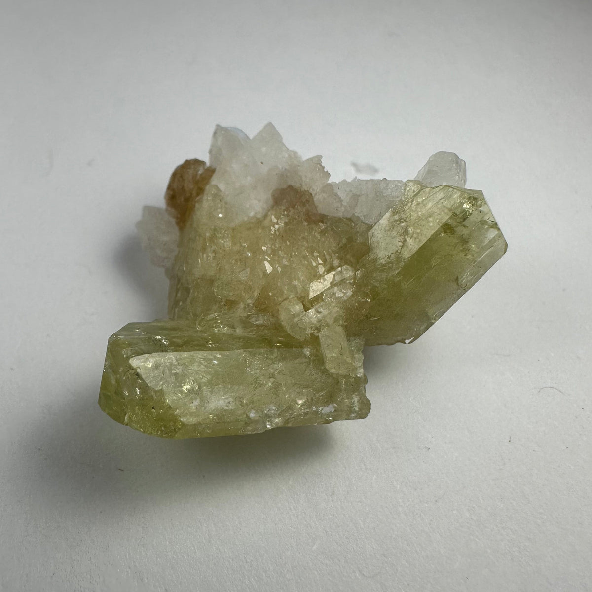 A cluster of Brazilianite crystals with pale yellow-green color growing on white Albite crystals, displayed on a light surface with subtle shadows.