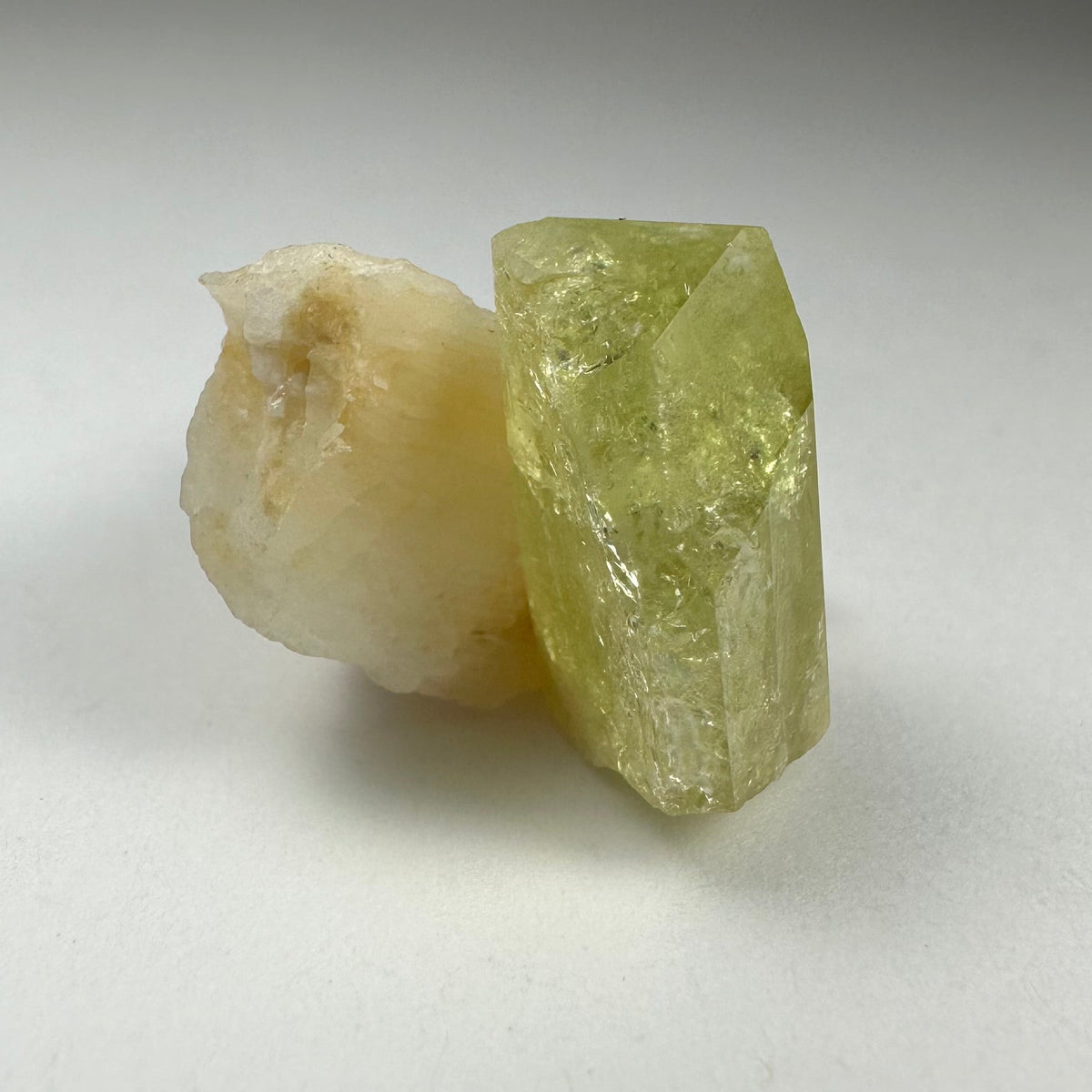 Translucent yellow-green Brazilianite crystal adjacent to an off-white, irregular Albite mineral on a plain white surface.