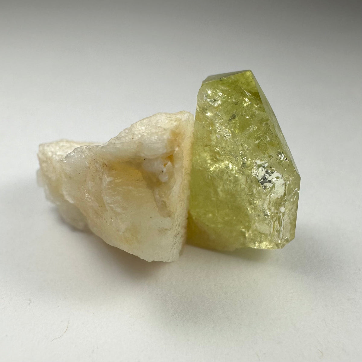 Yellow translucent Brazilianite crystal naturally attached to off-white Albite mineral against a light background.