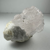 Translucent pink, naturally etched kunzite crystal resting on a rough, white and gray mineral base against a neutral background.