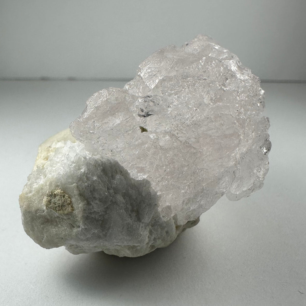 Translucent pink, naturally etched kunzite crystal resting on a rough, white and gray mineral base against a neutral background.