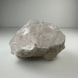 Translucent, textured kunzite crystal with irregular natural etchings resting on a rough, white mineral base against a soft gray background.