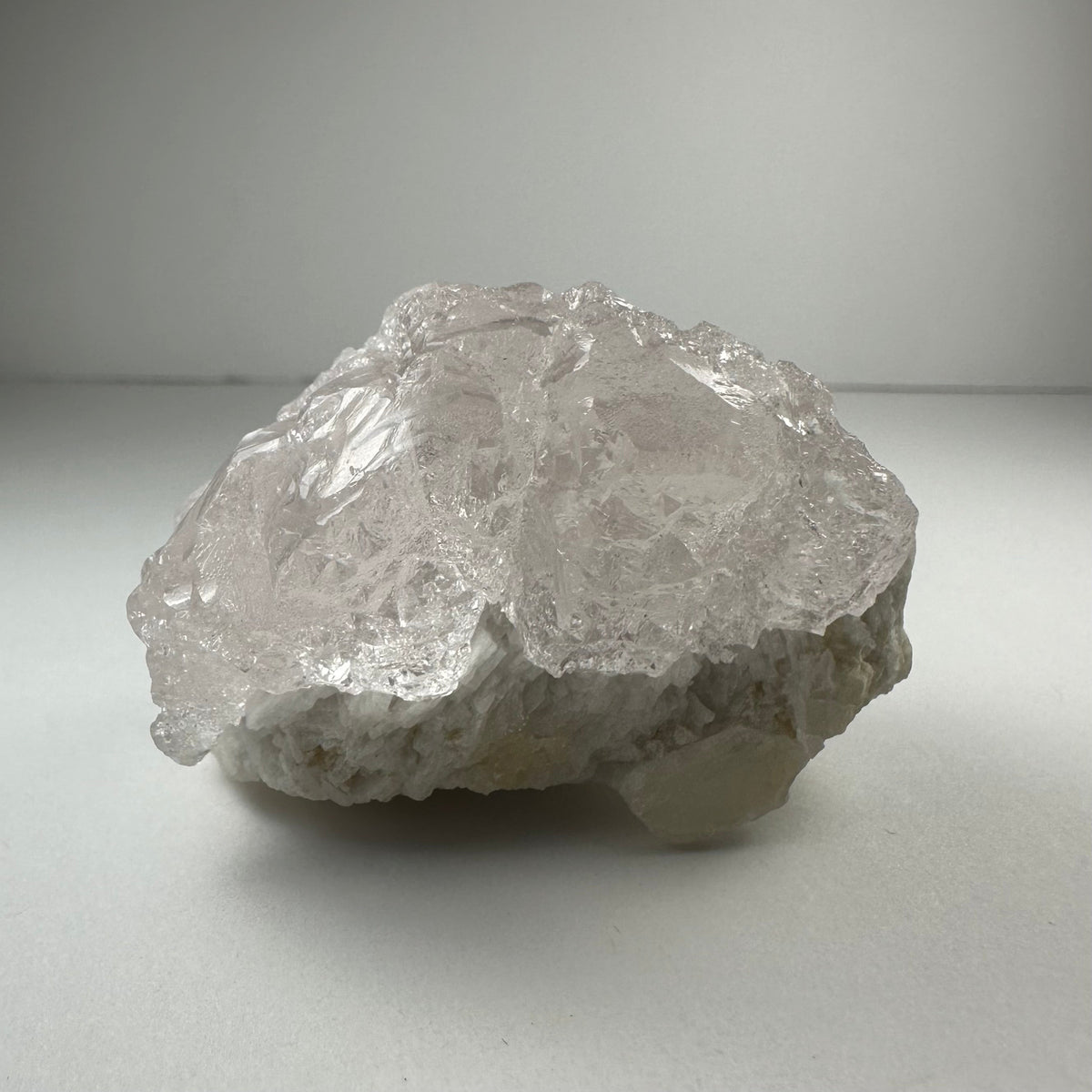 Translucent, textured kunzite crystal with irregular natural etchings resting on a rough, white mineral base against a soft gray background.