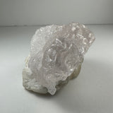Translucent, naturally etched kunzite crystal with a pale pink hue resting on a white mineral base against a neutral background.
