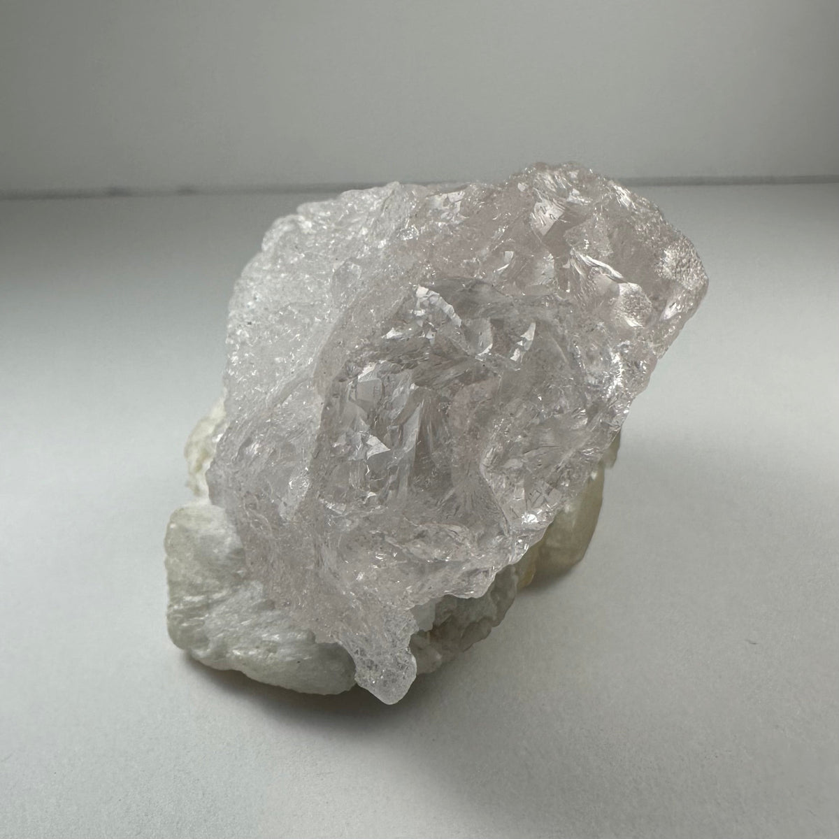 Translucent, naturally etched kunzite crystal with a pale pink hue resting on a white mineral base against a neutral background.