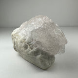 Translucent, naturally etched kunzite crystal with a rough, uneven surface resting on a matte white background, showing intricate internal textures and a white base matrix.