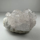Light pinkish natural kunzite crystal cluster with a rough, translucent surface and visible crystal facets, resting on a grayish base against a neutral background.