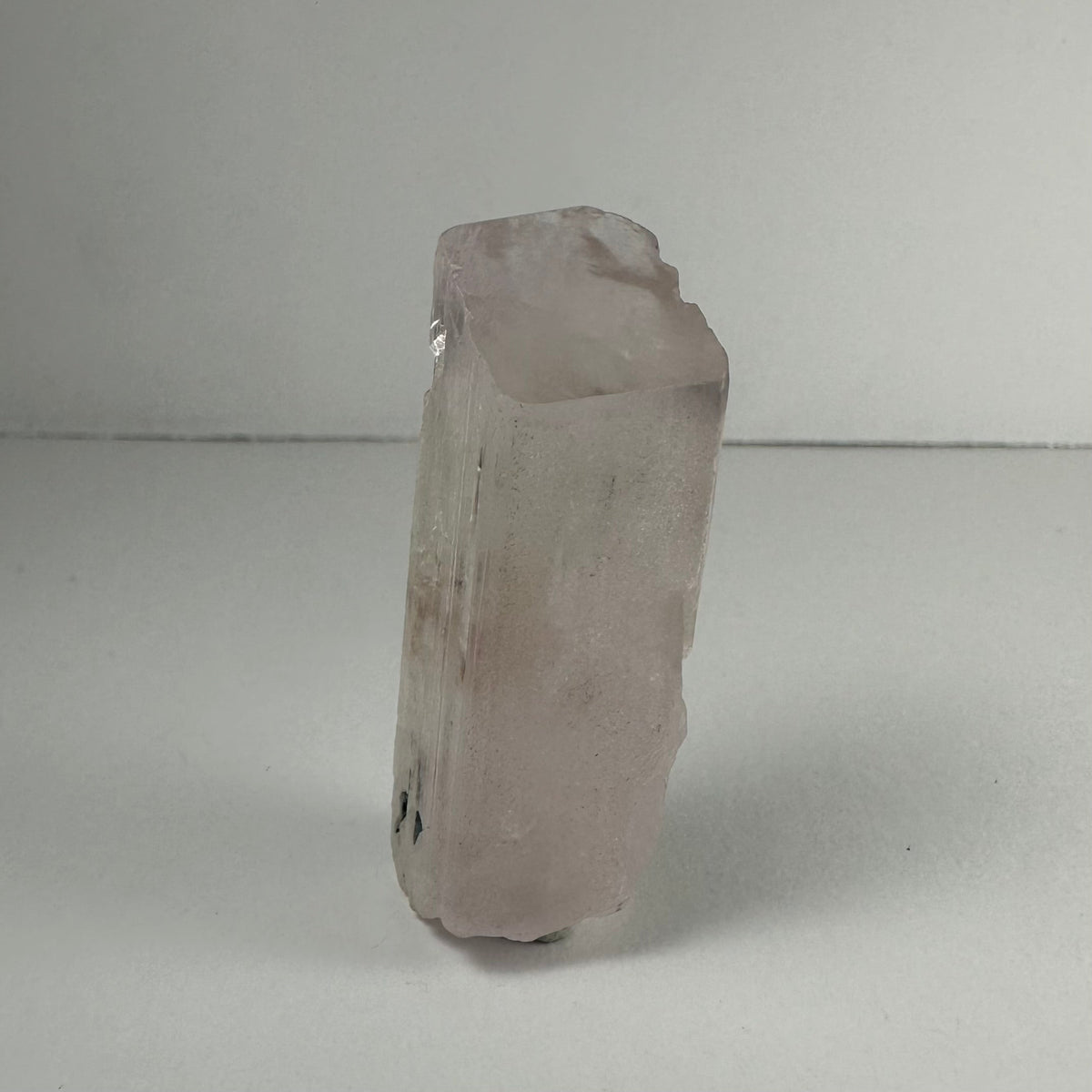 Translucent pale pink terminated spodumene kunzite crystal with a blocky shape standing upright on a white surface against a neutral background.