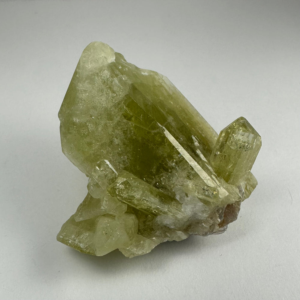 Cluster of translucent pale green Brazilianite crystals with elongated prismatic shapes and a rough white matrix base on a neutral background.