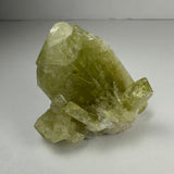 Translucent yellow-green Brazilianite crystal cluster with sharp geometric facets, displayed on a smooth light gray surface under soft lighting.