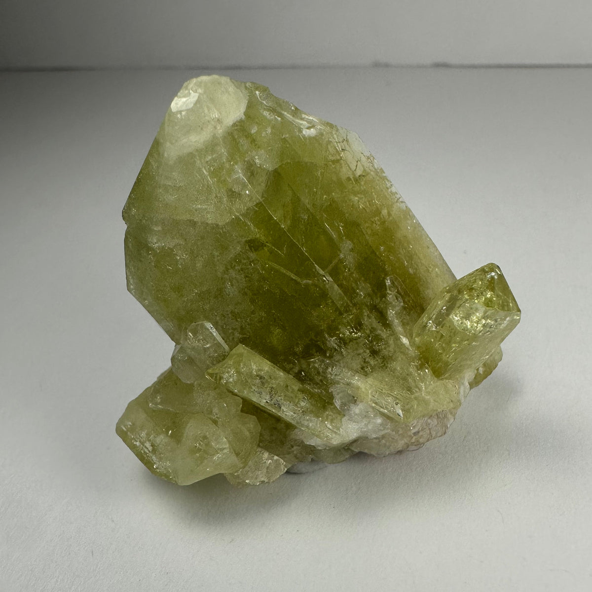 Translucent yellow-green Brazilianite crystal cluster with sharp geometric facets, displayed on a smooth light gray surface under soft lighting.