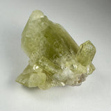 Translucent pale yellow-green Brazilianite crystal cluster with hexagonal prism shapes on a white background.