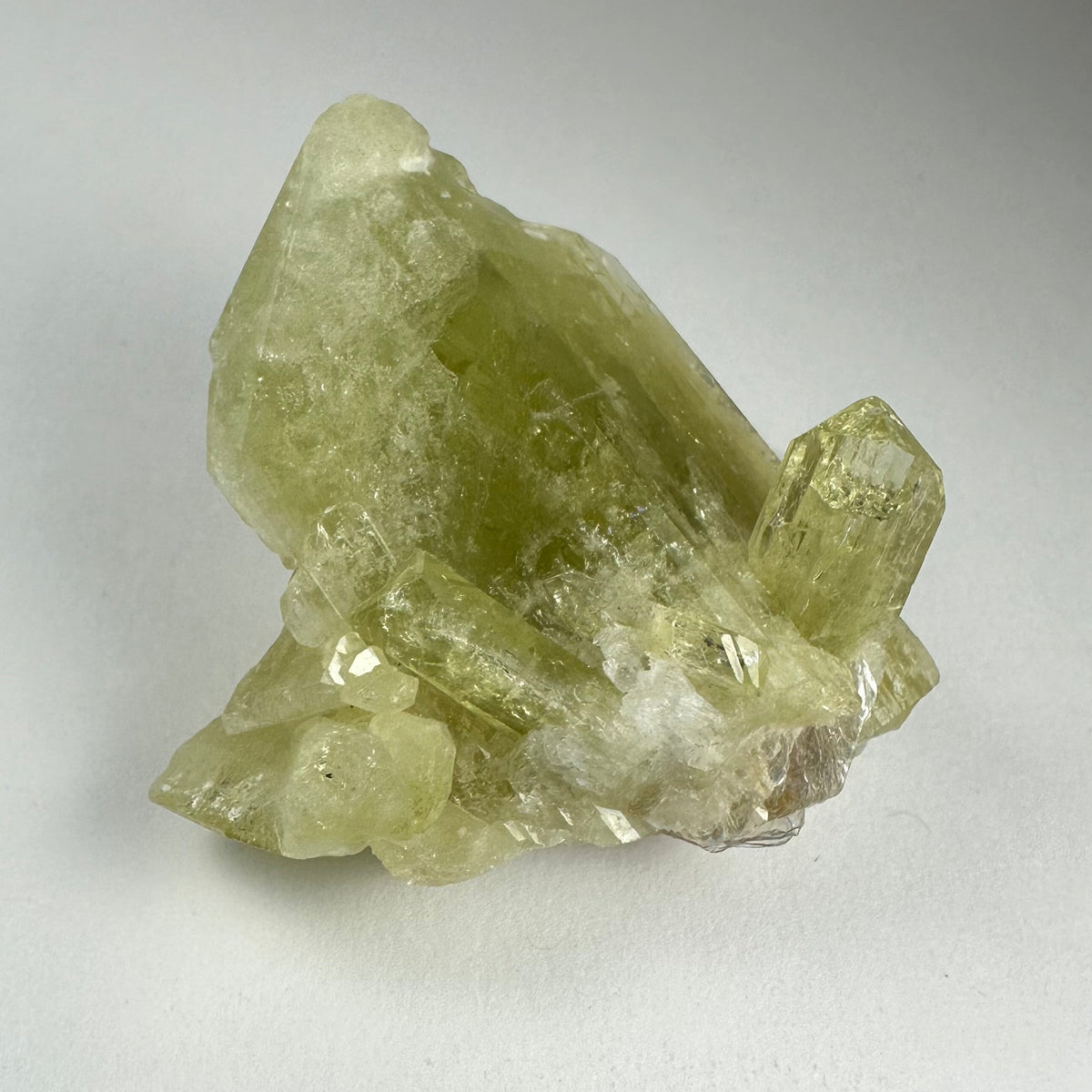 Translucent pale yellow-green Brazilianite crystal cluster with hexagonal prism shapes on a white background.