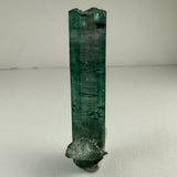 Tall slender indicolite tourmaline crystal with a rough greenish-blue surface and a small attached translucent mineral at its base, standing upright on a plain light background.