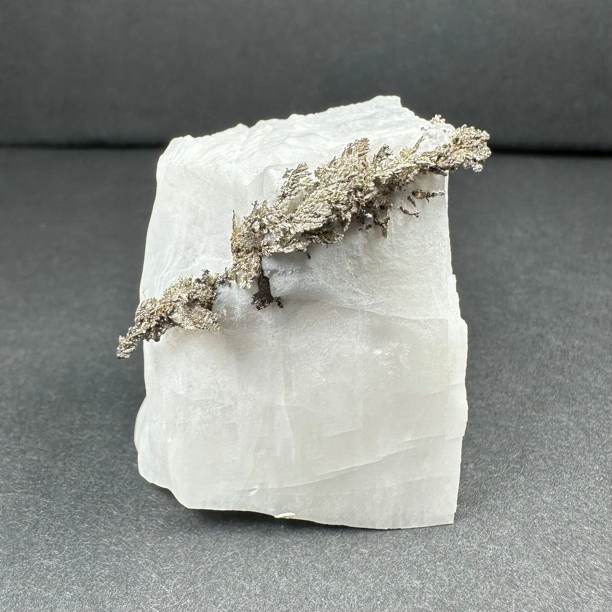 Chunk of white calcite with a jagged, silvery native silver dendritic formation extending across the top and side edges against a gray-black background.