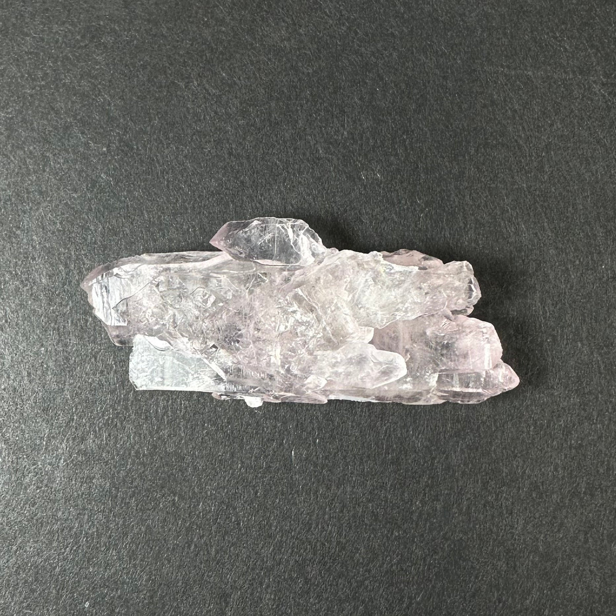 Translucent pale pink rose quartz crystal cluster with layered, jagged formations, displayed on a dark gray textured background.