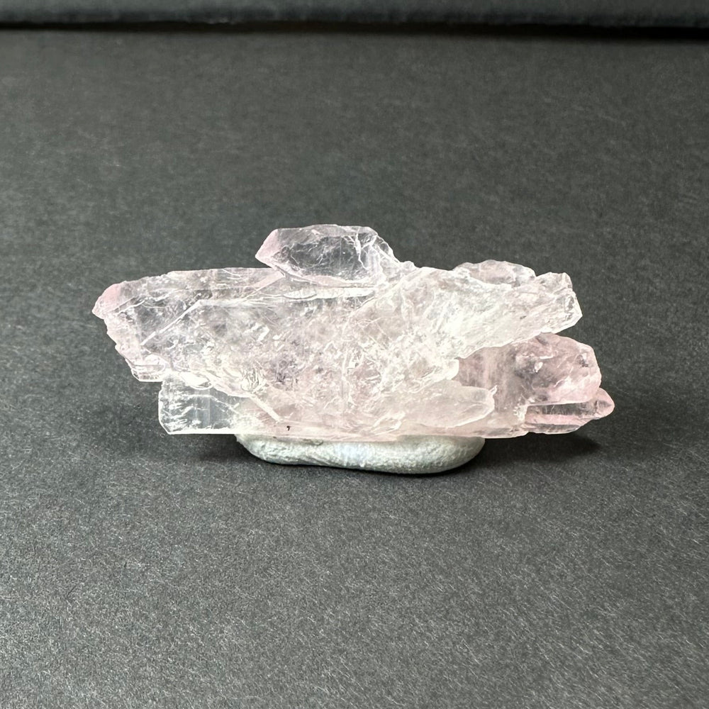 Translucent pale pink rose quartz cluster with jagged edges resting on a smooth, rounded gray stone against a dark background.