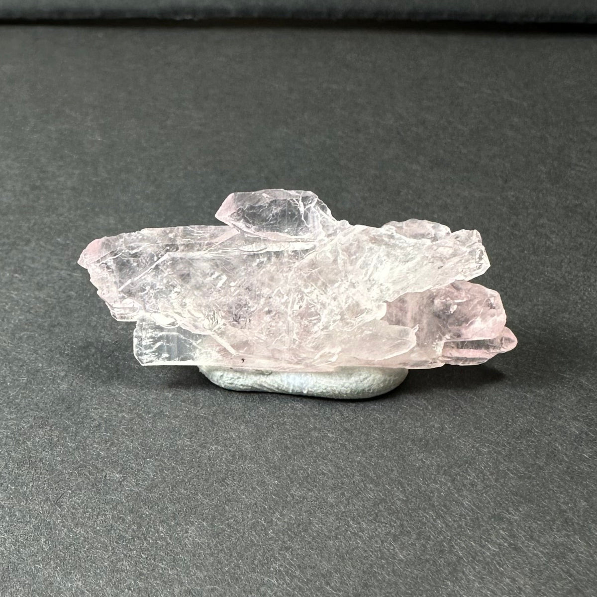 Translucent pale pink rose quartz cluster with jagged edges resting on a smooth, rounded gray stone against a dark background.