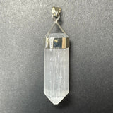 Clear selenite pendant with a pointed, elongated crystal held by a geometric sterling silver clasp on a textured gray background.